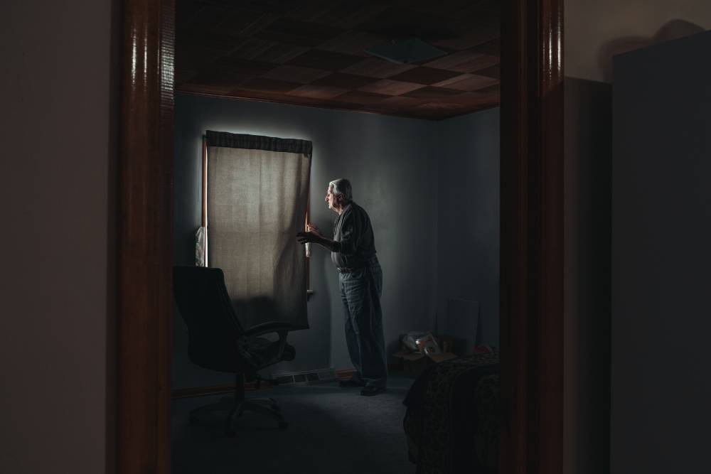 Elderly man in darkened room pulling back curtain to peer out